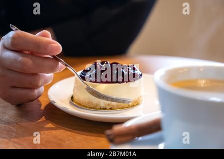 Die Hand einer Frau, die eine Gabel hält, um ein Stück Blaubeerkäse zu schneiden, um mit einer Kaffeetasse auf Holztisch zu essen Stockfoto