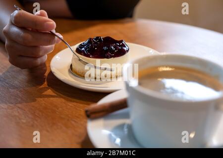 Die Hand einer Frau, die eine Gabel hält, um ein Stück Blaubeerkäse zu schneiden, um mit einer Kaffeetasse auf Holztisch zu essen Stockfoto