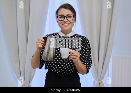 Erwachsene Hausfrau, die Kaffee zubereiten, Frauen halten Kaffeekanne und Tasse, stehen in der Nähe von Fenster mit Vorhängen, Pausenzeit zum Kaffeetrinken Stockfoto