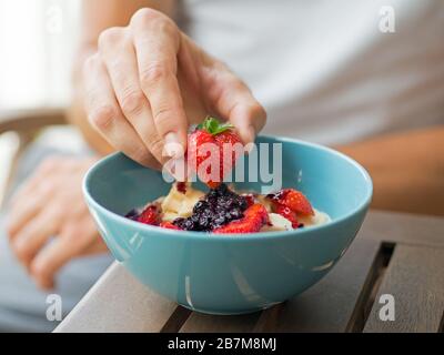 Herzförmige Erdbeere in Menschenhand und eine Frühstücksschale voller Obst mit veganem Joghurt. Hintergrund für gesundes Essen, Ernährung, Mahlzeiten, Nahaufnahme. Stockfoto