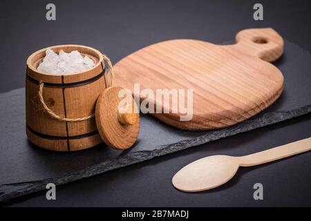 Kleines Holzfass zum Aufbewahren von Salz, Löffel und Schneidebrett mit Steinplatte in schwarzem Hintergrund. Draufsicht. Stockfoto