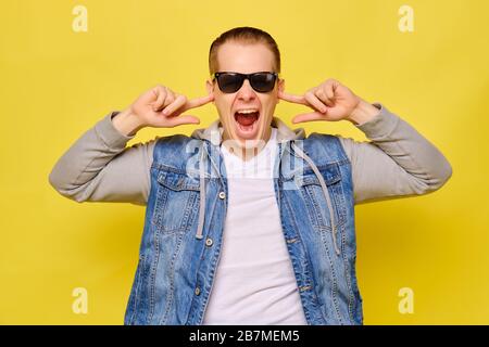 Stilvoller kaukasischer Mann in einer Jeans und Sonnenbrille auf gelbem Hintergrund. Nahaufnahme. Er bedeckt seine Ohren mit den Fingern seiner Hand. Stockfoto