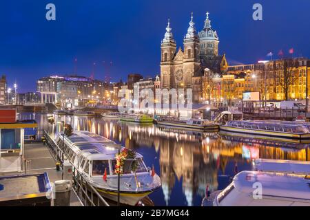 Amsterdam, Niederlande, Kanalszene in der Nacht mit der Basilika Sankt Nikolaus und Flussbooten.