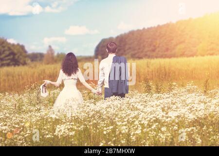 Ein junges glückliches Hochzeitspaar, das auf einem blühenden Feld spazieren geht und sich an einem sonnigen Sommertag die Hände hält. Eine Frau hält Schuhe in den Händen, ein Mann hält einen jac Stockfoto