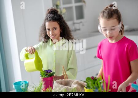 Dunkelhäutige Mädchen mit einer Gießkanne, eine fairhäutige Freundin sind in der Nähe. Stockfoto