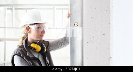 Frau Worker schaut auf die Wasserwaage und kontrolliert die Wand mit Hut, Brille und Ohrenschützer, weiße Wand mit Kopierraum Stockfoto