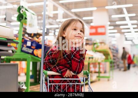 Ein fröhliches Mädchen hält einen Einkaufswagen und wählt Produkte nach Hause aus. Das Konzept des Einkaufs im Supermarkt, der Auswahl der Produkte, des Verkaufs. Stockfoto