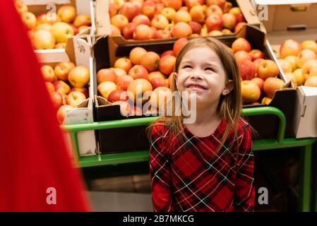 Ein kleines Mädchen von etwa 5 warf vor ihren Eltern ein Tantrum in einem Supermarkt. Das Kind schreit und schreit, bettelt Süßigkeiten von Mutter und Vater vor dem Hintergrund von Früchten und Äpfeln. Stockfoto