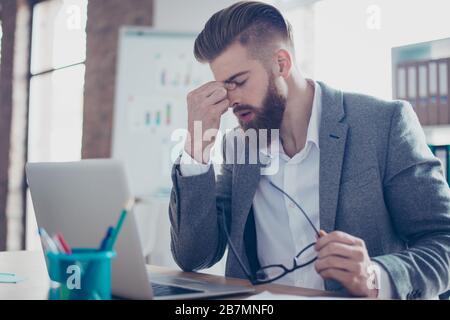 Nahaufnahme des müden bärtigen jungen Mannes mit starkem Migräne-Grimace. Er trägt die Schalenbekleidung, hält seine Brille und sitzt am Arbeitsplatz Stockfoto
