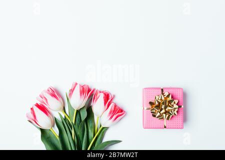 Kleiner rosafarbener Geschenkkasten neben einem Haufen frischer Tulpenblüten mit grünen Blättern auf weißem Grund, flacher Lay. Stockfoto