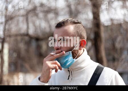 Ein Kerl in einer antiviralen Maske zieht aus und raucht auf der Straße eine Zigarette. Stockfoto