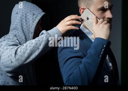 Zaporozhye. Ukraine. Februar 2020. Nahaufnahme eines jungen bärtigen Kerls in einem Business-Anzug mit einem iPhone XS max Ein Räuber in einer dunklen Kapuze, versuchen Sie es Stockfoto