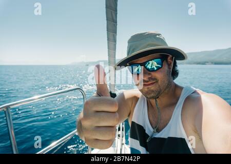 Mann mit Hut und Sonnenbrille, die am Bug eines segelschiffs den Daumen nach oben zeigt. Segel- und Segelkonzept. Stockfoto