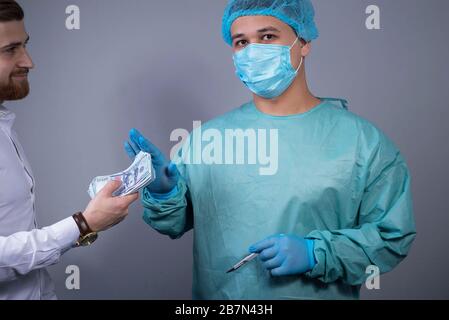 Porträt eines Chirurgen in einem sterilen Morgenmantel und Maske, mit Handschuhen, Blick auf die Kamera, und Verbot ihm, ihm eine Bestechung in Dollarscheinen zu überreichen. Stockfoto