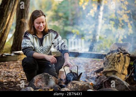 Mann und Frau, die auf dem Lagerfeuer im Wald am Ufer des Sees Kaffee zubereiten, Feuer machen und grillen. Glückliches Paar, das Finnland erkundet. Skandinavien Stockfoto