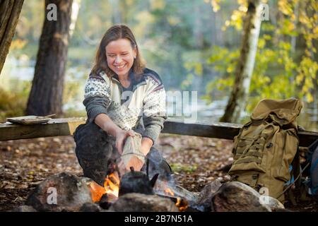 Mann und Frau, die auf dem Lagerfeuer im Wald am Ufer des Sees Kaffee zubereiten, Feuer machen und grillen. Glückliches Paar, das Finnland erkundet. Skandinavien Stockfoto