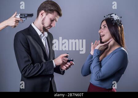 Studio-Porträt eines traurigen Kerls auf den Knien, der einem glücklichen Mädchen, das neben ihm steht, die Ehe vorschlägt, steht er einer Pistole von hinten gegenüber. Auf grauem Hintergrund Stockfoto