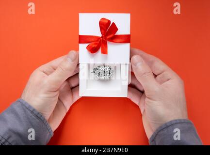 Männliche Hände halten eine halb geöffnete weiße Geschenkbox mit einem Metallmodell eines menschlichen Gehirns im Inneren und eine rote Schleife mit einem Band auf dem Deckel. Stockfoto