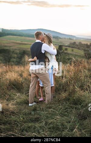 Fröhliche junge Familie, Vater, Mutter und zwei kleine Töchter, die im Freien Spaß haben und zusammen auf einem Feld spielen. Mutter und Vater küssen, Kinder sind es Stockfoto