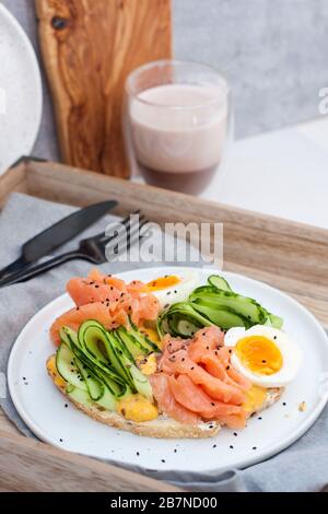 Sandwich mit Toastbrot, Räucherlachs, Frischkäse, geschnittener Gurke und gekochten Eiern auf weißem Teller. Stockfoto