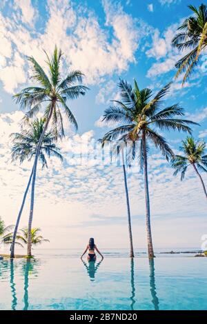 Eine junge Frau, die unter Palmen in einem Infinity-Pool steht Stockfoto