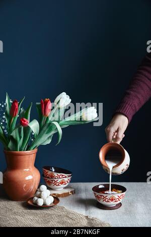 Kasachische Frau schüttet Milch in kasachischer Teeschale kese mit Schmuck in der Nähe von kurt und Tulpen auf dem Tisch während Nauryz-Festival auf dunkelblauem Hintergrund. Stockfoto