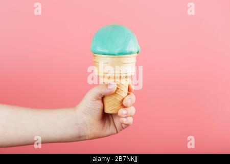 Eis in der Hand eines Kindes schmilzt auf einem rosafarbenen Hintergrund. Der Waffelkegel mit blauem Eis schmilzt. Stockfoto