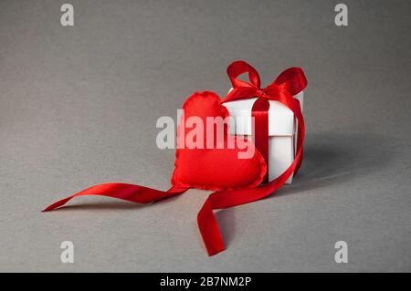 Konzept Valentinstag. Kleine Geschenkschachtel mit weicher roter Farbe, Herz und Weiß, mit roter Farbband auf grauem Hintergrund. Stockfoto