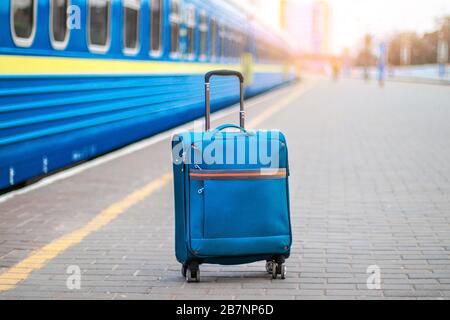 Blau tragen Koffer mit Rädern und ein Metallgriff steht auf der Perone der Station. Personenschnellzug im Hintergrund verschwommen. Reisekonzept Stockfoto