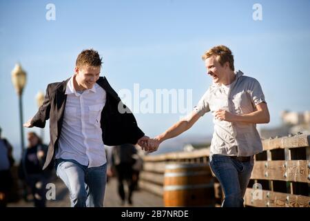 Zwei lächelnde junge homosexuelle Männer laufen zusammen, während sie die Hände halten. Stockfoto