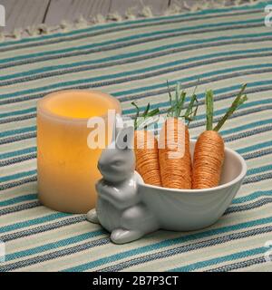 Leuchtende gelbe Wachskerze und keramische Osterhase Schüssel und künstliche Karotten Stockfoto