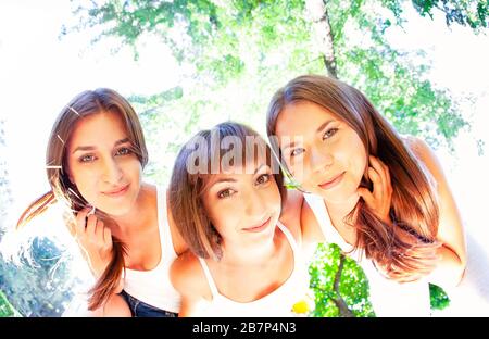 Von unten positive junge weibliche Freunde in legeren Outfits, die die Kamera umfassen und betrachten, während sie sonnigen Sommertag im Park verbringen Stockfoto