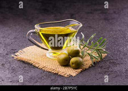 Olivenöl in einem durchsichtigen Glassauceboat, Rosmarin und Oliven auf schwarzem Grund. Nahaufnahme. Stockfoto