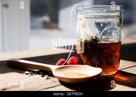 Hausgemachte Teebeutel, die in einem Einmachglas auf einem Holztisch vor einem Fenster, mit Tee, Himbeeren und einem Holzlöffel mit Honig gefüllt, eindringt. Stockfoto