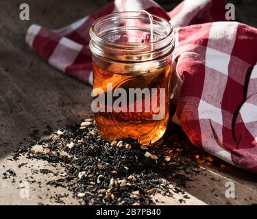 Hausgemachte Teebeutel, die in einem Einmachglas steppt, mit Tee und Himbeeren vor verschüttet, und ein rot-weiß kariertes Tuch hinter. Stockfoto