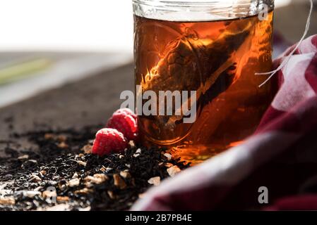 Hausgemachte Teebeutel, die in einem Einmachglas steppt, mit Tee und Himbeeren vor verschüttet, und ein rot-weiß kariertes Tuch hinter. Stockfoto