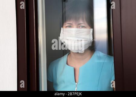 Coronavirus Quarantäne während der COVID-19-Epidemie. Frau in einer medizinischen Maske blickt aus dem Fenster Stockfoto