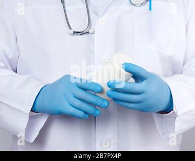 Der männliche Arzt in einem weißen, medizinischen Mantel und blauen Latexhandschuhen hält einen weißen Plastikbecher mit Pillen aus der Nähe Stockfoto