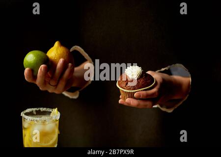 Hand in das Papierloch.Dunkler Hintergrund. Zitrone, Limette und Limonade. Erfrischung. Auf der anderen Seite ist ein Schokoladenkuchen. Die Wahl der richtigen Lebensmittel Stockfoto