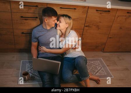 Oberansicht Foto eines kaukasischen Ehepaares jubelt in der Küche auf dem Boden zusammen, während er einen Kaffee trinkt und einen Computer benutzt Stockfoto