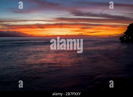 Weiches, gedämpftes Wasser reflektiert orange und violette Farben der Sonne, die am Horizont in der Küstenmeerküste untergeht. Stockfoto