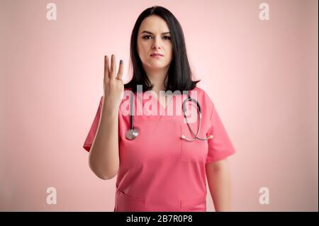 Porträt der schönen Frau Doktor mit Stethoskop mit rosa Schrubben, drei mit ihren Fingern auf einem rosa isolierten Hinterrund. Stockfoto