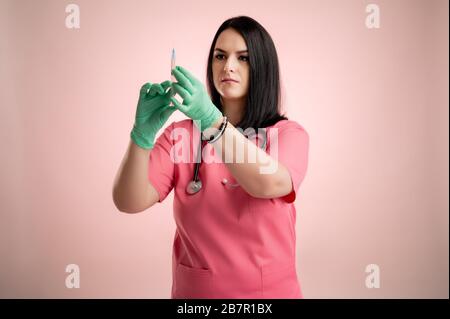 Portrait eines schönen Frauenarztes mit Stethoskop, das rosafarbene Schrubben trägt, mit medizinischem Handschuh und Spritze in der Hand, die auf einem rosafarbenen, isolierten Hinterrund posieren. Stockfoto