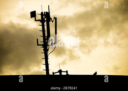 Silhouette einer Funk-Telekommunikationsnetzantenne, die auf einem Metallpol montiert ist und starke Signalwellen von der Oberseite des Dachs mit Sonnenaufgang an liefert Stockfoto