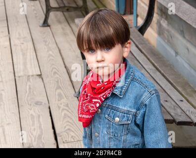 Liebenswürdiger, blauäugiger kleiner Junge mit einem niedlichen Pout, in einem Western-Outfit mit roter Bandana und blauer Jeans gekleidet. Stockfoto