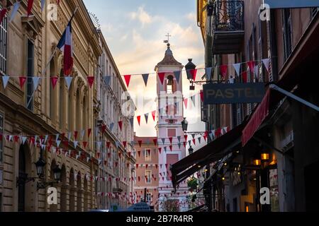 Die Flaggen sind über eine der Straßen in der Altstadt mit dem Uhrturm des Rusca Palace hinuntergedraftet, während die Sonne in Nizza, Frankreich, untergeht. Stockfoto