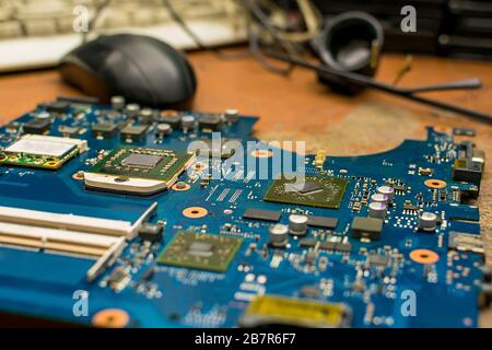 Die Hauptplatine des Computers liegt auf einem alten, aus Holz getragenen Reparaturtisch des Servicezentrums vor dem Hintergrund von Werkzeugen, einer Computermaus Stockfoto