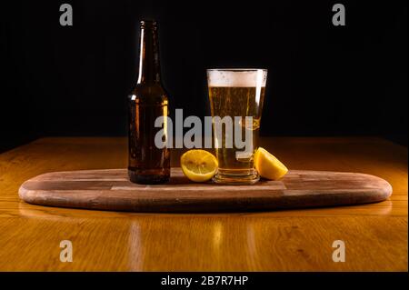 Isoliertes frisches Bier auf Holztisch mit Flasche und Zitronen, schwarzer Hintergrund Stockfoto