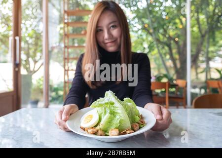 Eine schöne asiatische Frau, die im Restaurant einen Teller cäsar-salat auf dem Tisch hält und zeigt Stockfoto