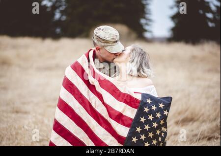 Shallow Focus Aufnahme eines amerikanischen Soldaten küsst seine liebevolle Frau, während sie in eine amerikanische Flagge gehüllt ist Stockfoto